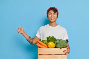 Joyful farmer guy holding box with fresh vegetables in his hand, shows thumb up, wearing white...