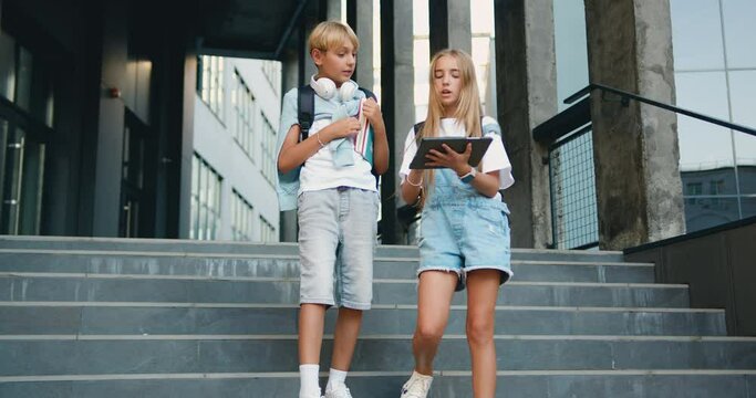 Two teenage students boy and girl friends using digital tablet computer exchange lectures near school building summer day on stairs outside campus. Friendship. Back to school.