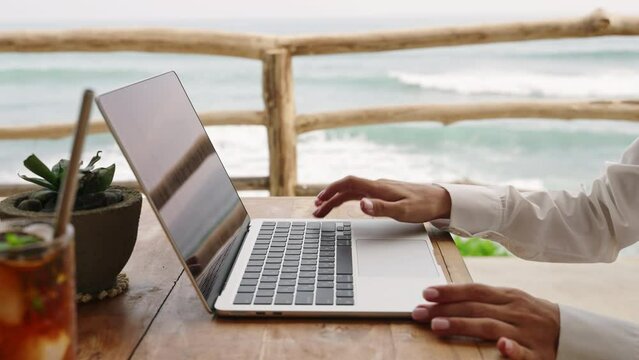 Blonde woman starts working day, opens laptop lid in tropical cafe with ocean view. Female begins typing on keyboard sitting in authentic restaurant. Business person turns on computer for meeting.