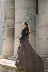 Woman in black dress stands on steps near large columns of ancient temple at sunset. Model girl in nature autumn day. Female near ancient Baroque Roman Catholic church in Pidhirtsi Lviv region Ukraine