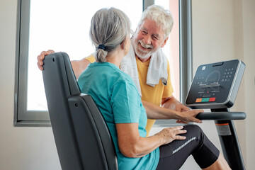 Athletic happy senior couple together in gym area, woman doing exercise bike while talks with a smiling elderly man