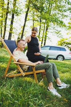 A happy man and his pregnant wife are working on a laptop while sitting in nature. A young couple is relaxing in nature and watching a movie on a laptop.