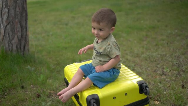 A little boy sits on a yellow suitcase in nature. 