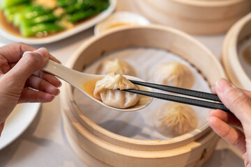Fototapeta na wymiar Person taking a dumpling from spoon with chopsticks. Closed up freshly steamed baozi dumplings in white spoon with bamboo steamer background.