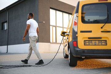 A african american man stands next to yellow electric delivery van at electric vehicle charging...