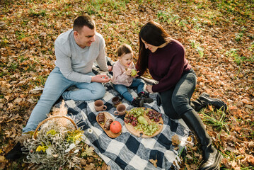 Naklejka na ściany i meble Autumn mood top view. Mother, father, kid have picnic in autumn forest in nature. Mom, dad hugs daughter child sitting on blanket on yellow leaves in park. Family spending time together at sunset.