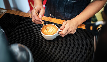 Close-up of the hand of a barista with a wooden spoon making or preparing coffee foam in a cup of...