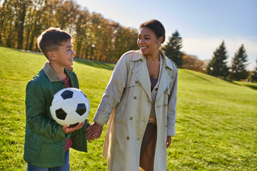 happy african american boy holding soccer ball, walking with mom on green field, outerwear, autumn © LIGHTFIELD STUDIOS