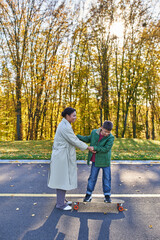 Fototapeta premium mother and son in park, happy african american woman holding hands with boy on penny board, autumn