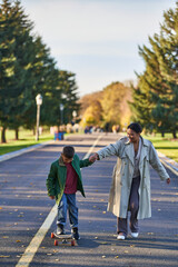 boy riding penny board and holding hands with mother, african american, golden leaves on asphalt