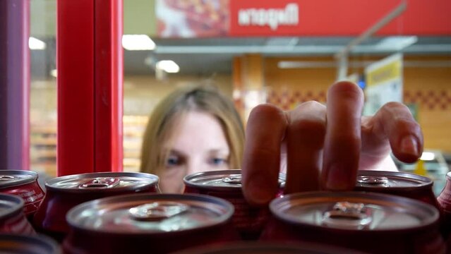 A young lady slides the door of a store fridge and takes one can of drink close-up