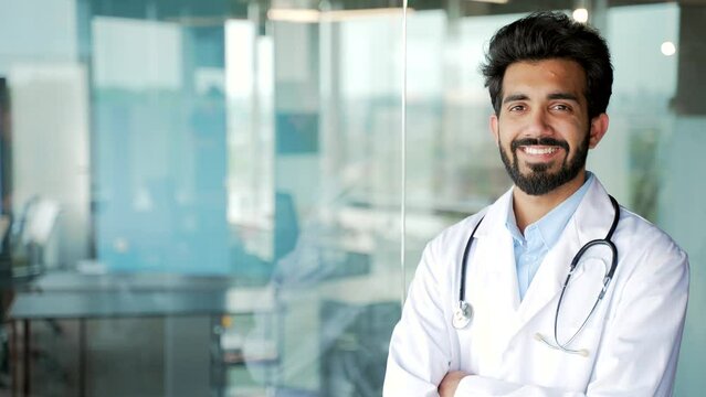 Portrait of a young smiling bearded medical doctor in white coat looking at camera in modern hospital clinic. Positive medical worker physician posing in office. Template for advertising. Copy space