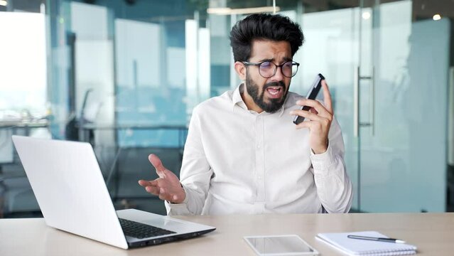 Angry excited young businessman arguing talking on smartphone while sitting at workplace at desk in modern office. Annoyed boss shouting into the phone gesturing, describing a problem to be solved