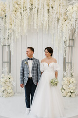 Newlyweds with a bouquet,  standing on wedding ceremony under arch decorated white flowers. Bride and groom get married. Full length of attractive woman wearing dress and man while ceremony. Marriage.