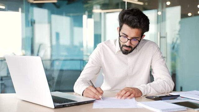 Busy young man financier making financial calculations using a calculator, typing on laptop, taking notes while sitting at workplace. Confident male accountant in glasses working in modern office
