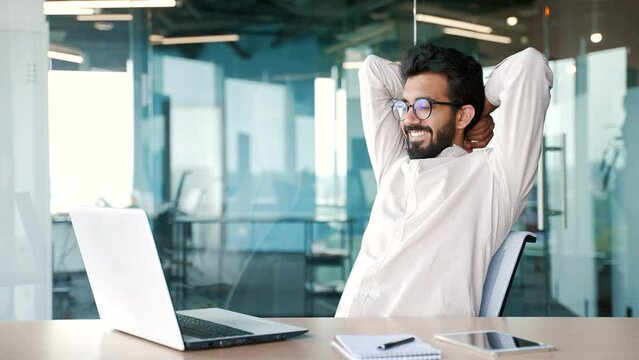 Young Bearded Businessman Wearing Glasses Finished Work On Laptop While Sitting At Workplace In Modern Office. Happy Handsome Male Stretches In A Chair, Throwing His Hands Behind His Head. Work Done