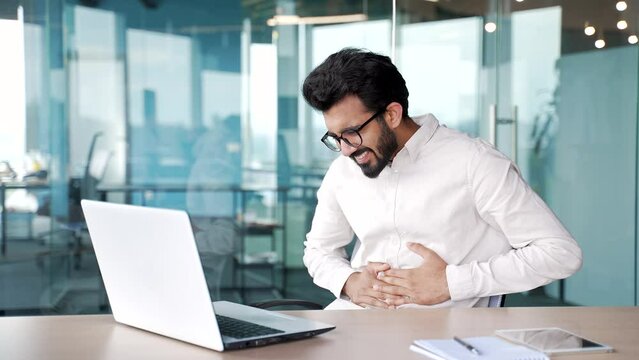 Young businessman feels stomach pain while working on a laptop while sitting at workplace in modern office. Sick worker has heartburn, gastritis or poisoning. Male suffers from spasms and constipated