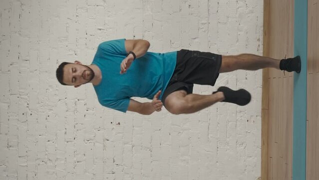 Vertical video. Man fitness coach in sportswear doing high knee jums in place warm up for online class at the home studio.
