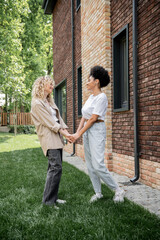 Fototapeta premium side view of delighted multiethnic lesbian couple holding hands near new private cottage