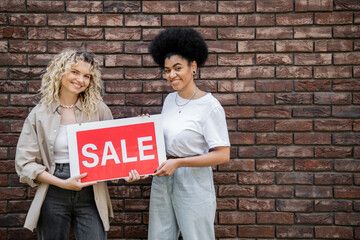 cheerful multiethnic lesbian couple holding sale signboard and looking at camera near cottage