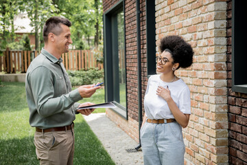 real estate agent with folder and pen talking to smiling african american client near new cottage