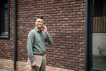 Fototapeta na wymiar successful real estate agent holding folder and talking on mobile phone near building on street