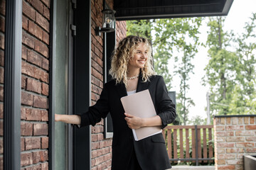 successful and happy real estate agent holding folder and looking away near door of new house