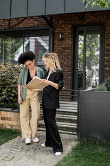blonde real estate agent showing documents to african american client near modern house for sale