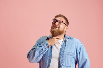 Bearded man in casual clothes and glasses standing with thoughtful, dreamy face against pink studio...