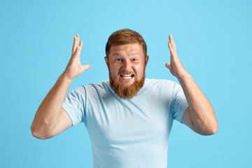 Bearded redhead man emotionally shouting, expressing anger and irritation against blue studio...