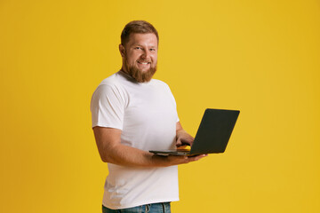 Smiling, positive, bearded man standing with laptop, working, shopping against yellow background. Concept of emotions, lifestyle, business and freelance, job fair, communication, ad. Copy space for ad