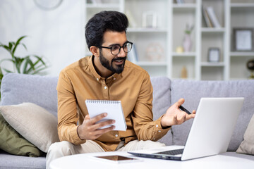 Young hispanic male student studying at home sitting on sofa, watching online course using laptop for video call, writing data in notebook, happy in living room
