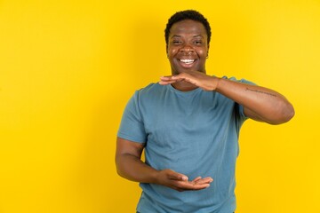 Young handsome man standing over yellow studio background gesturing with hands showing big and large size sign, measure symbol. Smiling looking at the camera. - obrazy, fototapety, plakaty
