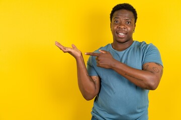 Young handsome man standing over yellow studio background pointing and holding hand showing adverts - obrazy, fototapety, plakaty