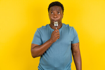Very hungry Young handsome man standing over yellow studio background holding spoon into mouth...