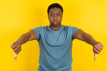 Young handsome man standing over yellow studio background being upset showing thumb down with two...