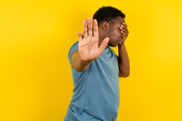 Young handsome man standing over yellow studio background covers eyes with palm and doing stop...