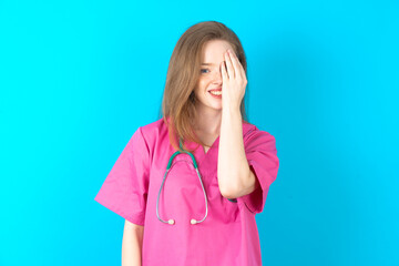 beautiful doctor woman standing over blue studio background  covering one eye with her hand,...