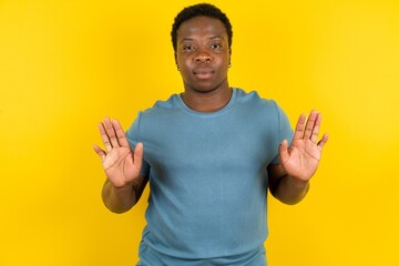 Serious Young handsome man standing over yellow studio background pulls palms towards camera, makes...