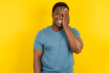 Young handsome man standing over yellow studio background covering one eye with her hand, confident...