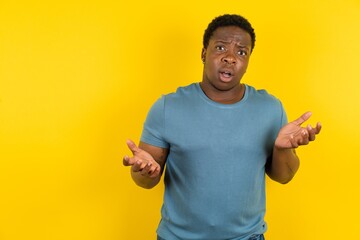 Frustrated handsome African American standing over yellow studio background feels puzzled and hesitant, shrugs shoulders in bewilderment, keeps mouth widely opened, doesn't know what to do.