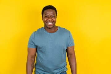 handsome African American standing over yellow studio background keeps teeth clenched, frowns face in dissatisfaction, irritated because of much duties.