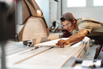 Young carpenter cutting a piece of wood in using a circular saw in furniture factory - obrazy, fototapety, plakaty
