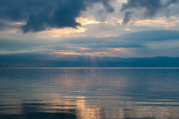 The sun setting behind mountains in Croatia. The Mediterranean Sea is calm and clear, reflecting the sunbeams. The sky is orange and covered with clouds. Many cities along the coastal line. Calmness