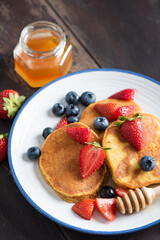 Pancakes with strawberries, blueberries and honey on plate, brown wooden table background. Closeup view