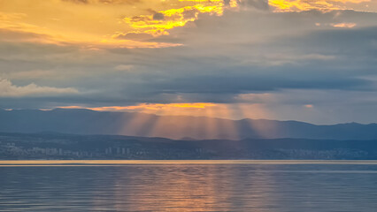 The sun setting behind mountains in Croatia. The Mediterranean Sea is calm and clear, reflecting the sunbeams. The sky is orange and covered with clouds. Many cities along the coastal line. Calmness