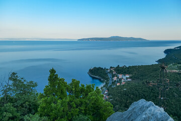 A small metal cross on a rock with a panoramic view of the shore along Medveja, Croatia seen from above. The town is surrounded by thick forest. Endless horizon. An island in the back. Summer holidays