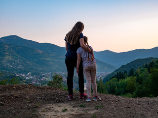 Fototapeta premium Travelers mother and daughter standing back view beautiful Carpathian Mountains background on sunset, having fun spending time together enjoying nature. Active lifestyle family travel tourism concept.