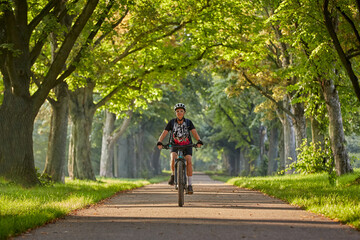 attractive senior woman cycling with her electric mountain bike in a beautiful old oak tree and chestnut avenue in Ludwigsburg, Baden-Wuerttemberg, Germany
