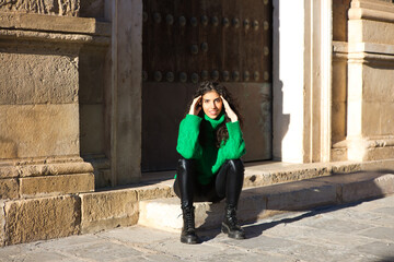 Fototapeta na wymiar pretty young brunette woman with curly hair and green woollen coat is sitting on a step at the entrance of a building in seville, spain. The woman is happy and enjoying her holiday in the city.